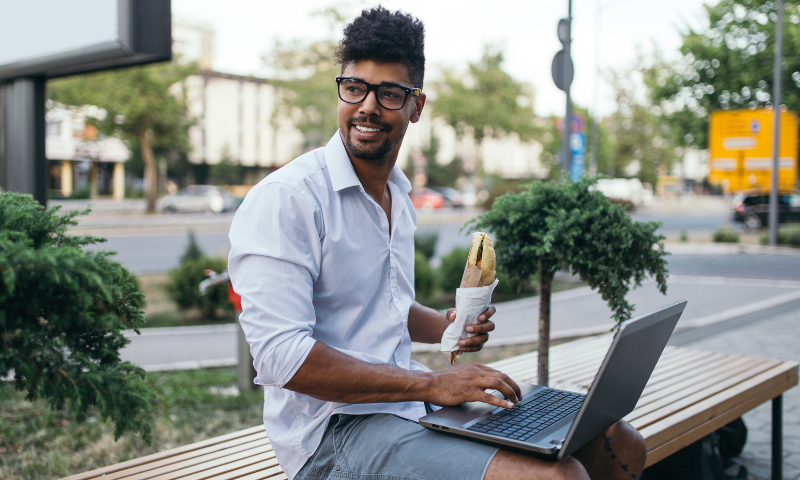 Young man eating a sandwich on a bench outside with his laptop on his lap