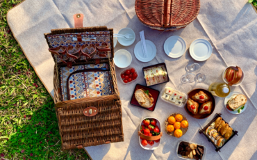 A picnic basket layed out on a blanket with food.