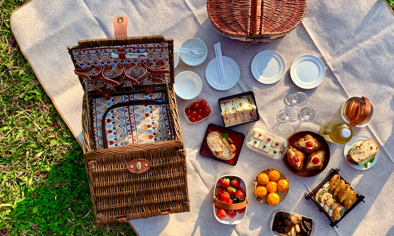 A picnic basket layed out on a blanket with food.